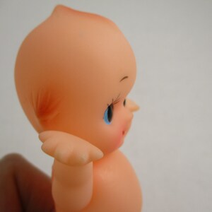 May include: A vintage Kewpie doll in a side profile view. The doll is peach-colored with blue eyes, long eyelashes, and rosy cheeks. It has a small, upturned nose and a tuft of reddish-orange hair.