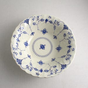 May include: A white ceramic bowl with a scalloped edge and a blue floral pattern. The bowl features a central floral design and delicate blue vines and flowers around the rim. The overall design is symmetrical and elegant.