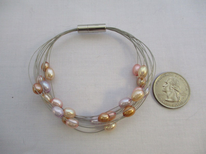 Op de afbeelding: Een zilveren draad armband met een magnetische sluiting en meerdere strengen parels in tinten roze, perzik en lavendel.