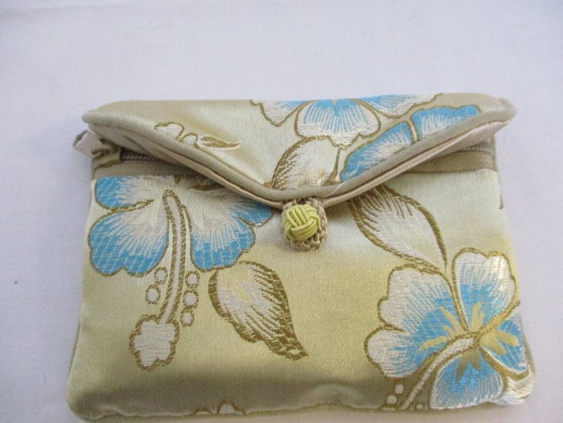 Op de afbeelding: Een kleine, gouden clutch met een bloemenpatroon. De clutch heeft een blauw en wit bloemenpatroon met een gele knoopsluiting.