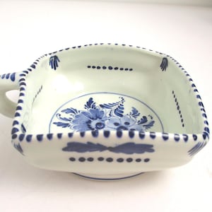 May include: A square ceramic bowl with a single handle, featuring a white base with hand-painted blue floral motifs in the center and blue dotted line patterns along the rim and sides. The bowl has a rounded bottom and a flared top edge.