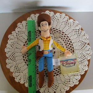 Puede incluir: Mu&ntilde;eco de peluche de Woody de Disney Store, con sombrero marr&oacute;n, camisa amarilla, chaleco, pantalones azules y botas marrones. Una regla verde est&aacute; al lado del mu&ntilde;eco. El mu&ntilde;eco mide unos 30 cm de alto.