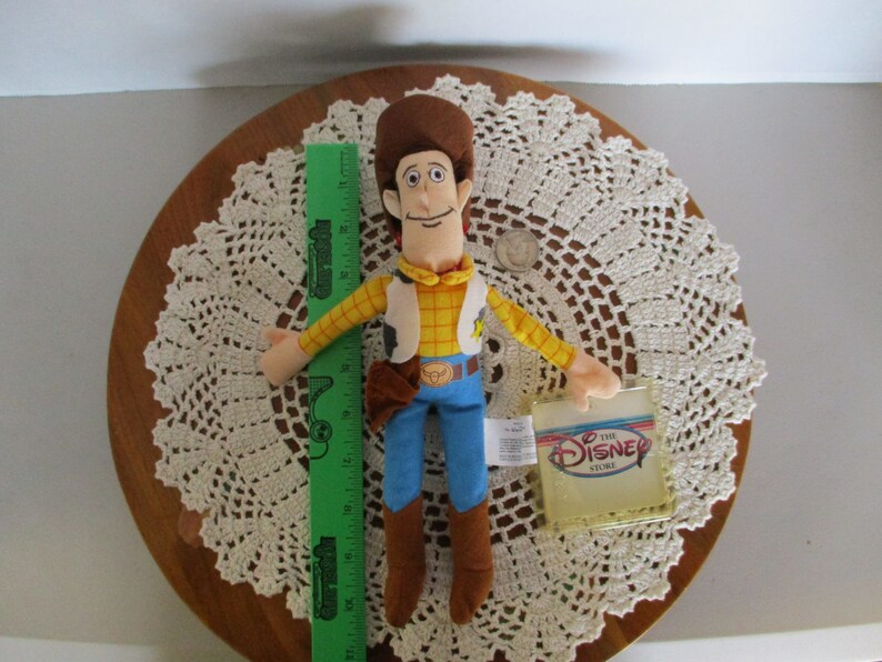 Puede incluir: Mu&ntilde;eco de peluche de Woody de Disney Store, con sombrero marr&oacute;n, camisa amarilla, chaleco, pantalones azules y botas marrones. Una regla verde est&aacute; al lado del mu&ntilde;eco. El mu&ntilde;eco mide unos 30 cm de alto.
