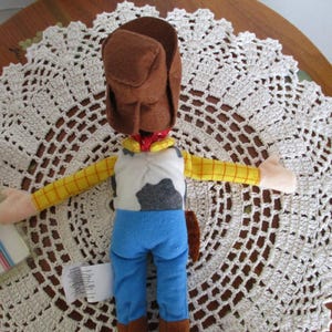 Puede incluir: Un peluche de un personaje de vaquero, con sombrero marr&oacute;n, mangas amarillas a cuadros, chaleco blanco con estampado de piel de vaca negra y gris, pantalones azules y pa&ntilde;uelo rojo. El juguete est&aacute; sobre un tapete de encaje blanco.