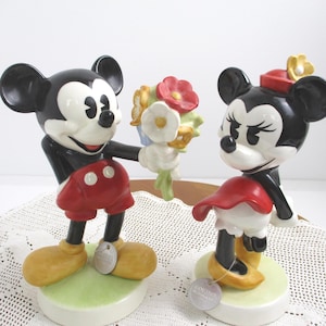 May include: Two ceramic figurines of Mickey and Minnie Mouse. Mickey, in red shorts, offers a bouquet to Minnie, who wears a red dress and hat. Both figures have black ears and are on green and white bases.