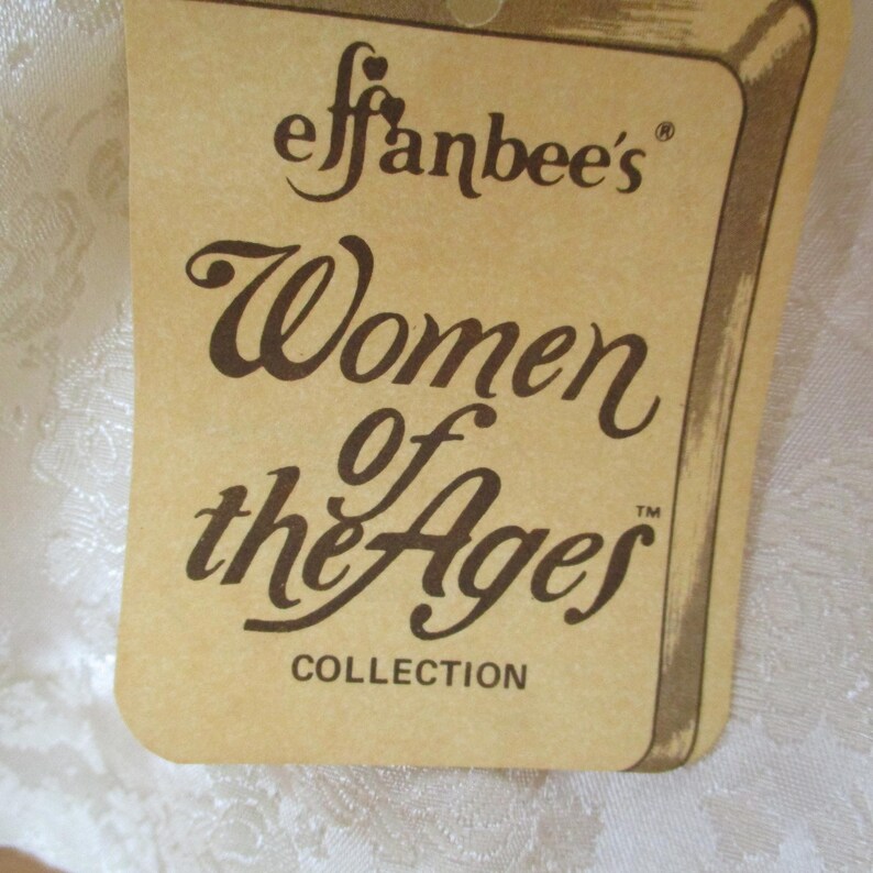 Puede incluir: Una etiqueta de papel marr&oacute;n con el texto "Effanbee's Women of the Ages Collection" en letras marrones.