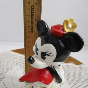 May include: A vintage Minnie Mouse figurine, featuring a black and white face with closed eyes, a red hat, and a yellow flower. The figurine is wearing a red dress and has black ears. A wooden ruler is in the background.