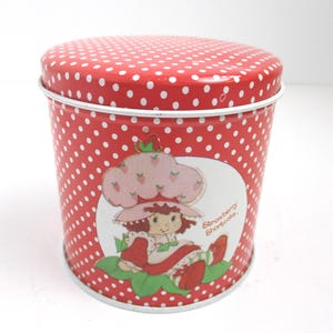 May include: A cylindrical red metal tin with a white polka dot pattern and a removable lid. The front features a circular graphic of the character Strawberry Shortcake with the text Strawberry Shortcake printed in orange script to the right.