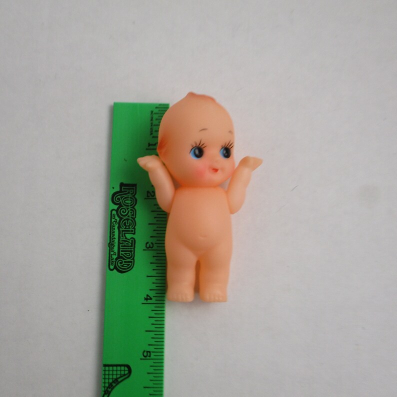May include: A small, nude doll with a peach-colored skin tone, blue eyes, and a slight smile. The doll is standing next to a green ruler, which measures the doll's height. The doll's arms are raised, and it has a small amount of red hair.