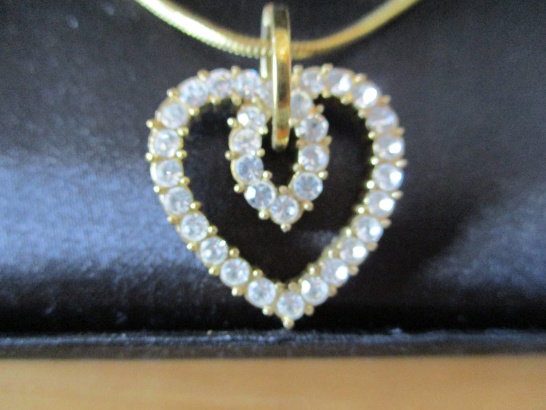 Puede incluir: Un collar de cadena dorada con un colgante de coraz&oacute;n doble. El colgante est&aacute; hecho de diamantes de imitaci&oacute;n transparentes.