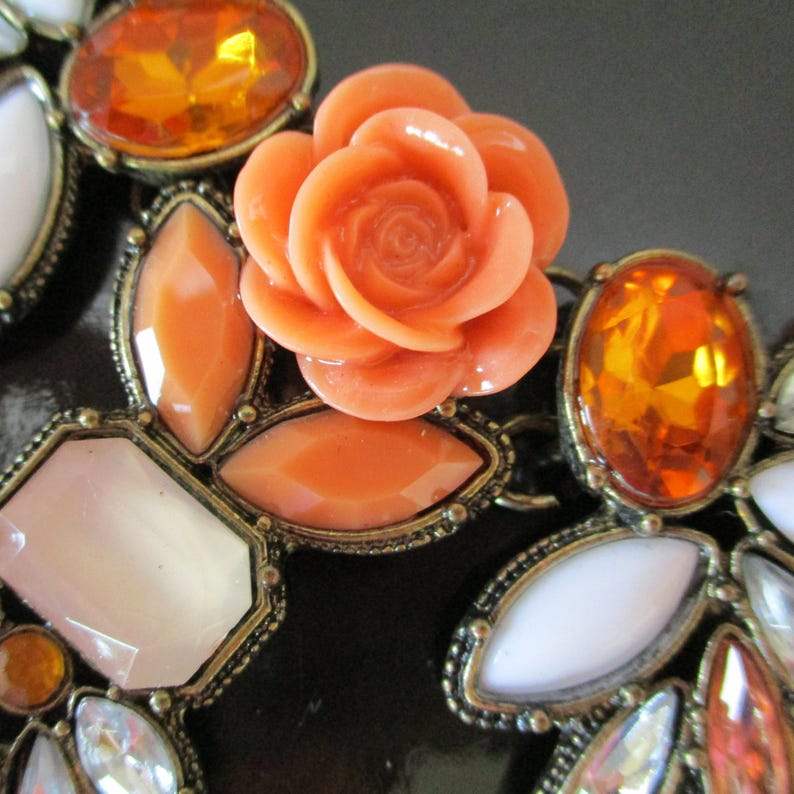May include: A close-up of a necklace with a coral-colored rose charm and various orange, white, and clear rhinestones.