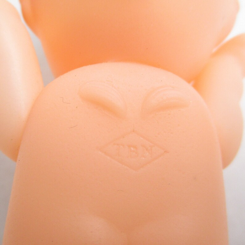 May include: A close-up of a vintage, peach-colored baby doll. The doll's back is visible, with a diamond-shaped mark that reads "TBN". The doll's arms and head are also visible.