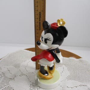 May include: A ceramic Minnie Mouse figurine. The figure is black, white, and red, with a yellow flower on her hat and yellow shoes. The figurine is standing on a light green base.
