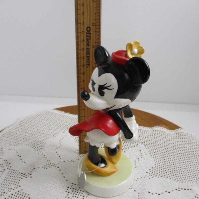 May include: A ceramic Minnie Mouse figurine. The figure is black, white, and red, with a yellow flower on her hat and yellow shoes. The figurine is standing on a light green base.