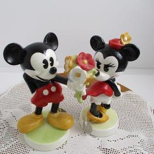 May include: A pair of ceramic figurines depicting Mickey and Minnie Mouse. Mickey, in red shorts and yellow shoes, offers a bouquet to Minnie, who wears a red dress and hat. Both figures have black ears and are on green and white bases.