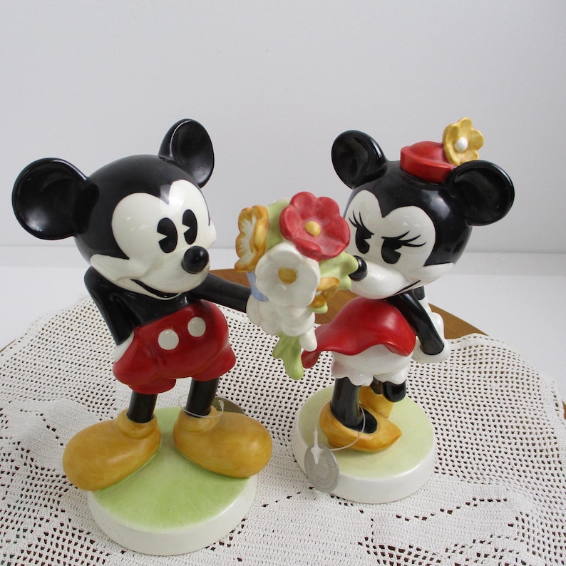 May include: A pair of ceramic figurines depicting Mickey and Minnie Mouse. Mickey, in red shorts and yellow shoes, offers a bouquet to Minnie, who wears a red dress and hat. Both figures have black ears and are on green and white bases.