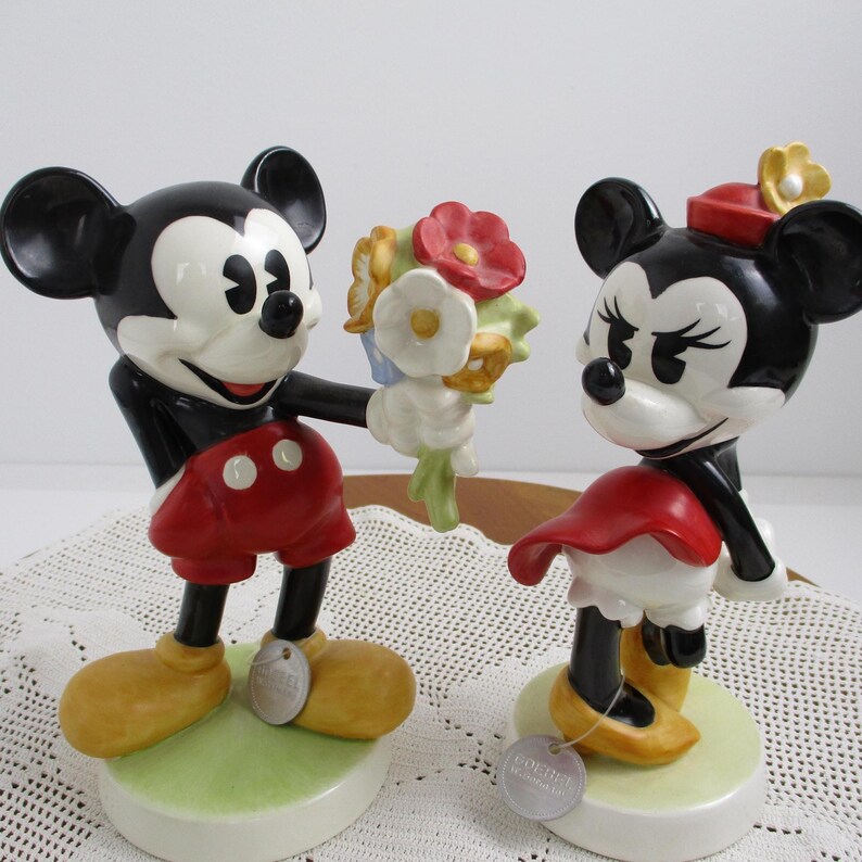 May include: A pair of ceramic figurines featuring Mickey Mouse and Minnie Mouse. Mickey, in red shorts, offers a bouquet to Minnie, who wears a red dress and hat. Both figures have black ears and are on green and white bases.