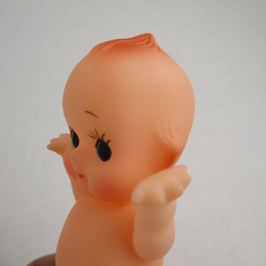 May include: A vintage rubber baby doll with a peach skin tone. The doll has large black eyes, long eyelashes, and rosy cheeks. Its hair is styled with a small curl on top. The doll's arms are outstretched, and it has a small mouth.