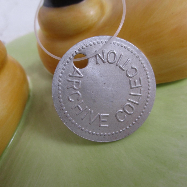 May include: Close-up of a silver, circular tag with the words "ARCHIVE COLLECTION" embossed around the edge. The tag is attached to a yellow object with a clear string. The background is a light green color.
