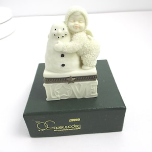 May include: A cream-colored figurine of a child in a textured snowsuit hugging a snowman. The base is a rectangular box with the word LOVE embossed on the front, featuring a small star and a dark metal hinge. The box sits on a dark green base.