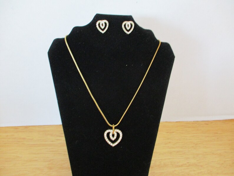 Puede incluir: Un collar de cadena dorada con un colgante de coraz&oacute;n doble y pendientes a juego. Los corazones est&aacute;n engastados con diamantes de imitaci&oacute;n transparentes.