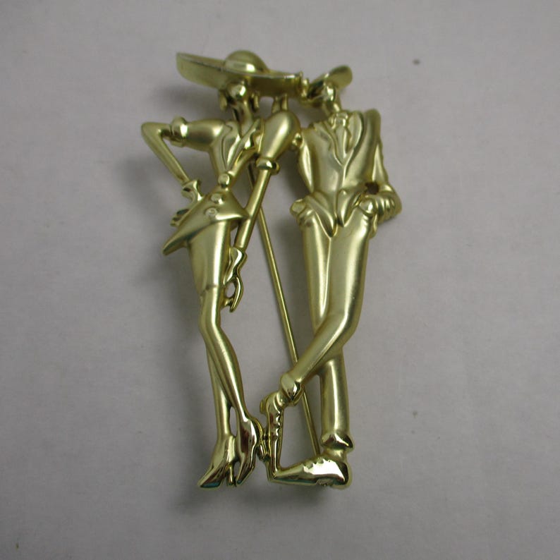 May include: Gold-toned metal brooch depicting a couple dancing. The woman is wearing a hat and the man is wearing a suit. The brooch is in a vintage style.
