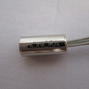 Op de afbeelding: Een zilveren sleutelhanger met een zilveren kabel en een cilindrische hanger gegraveerd met het woord "HONORA".
