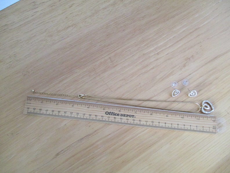 Puede incluir: Un conjunto de collar y pendientes de oro con un colgante en forma de coraz&oacute;n y pendientes a juego. El colgante y los pendientes est&aacute;n adornados con brillantes transparentes.