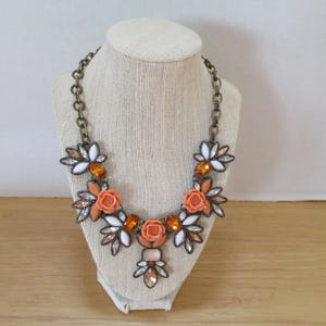 May include: A statement necklace with a floral design featuring orange and white flowers, clear and amber rhinestones, and a gold-tone chain.