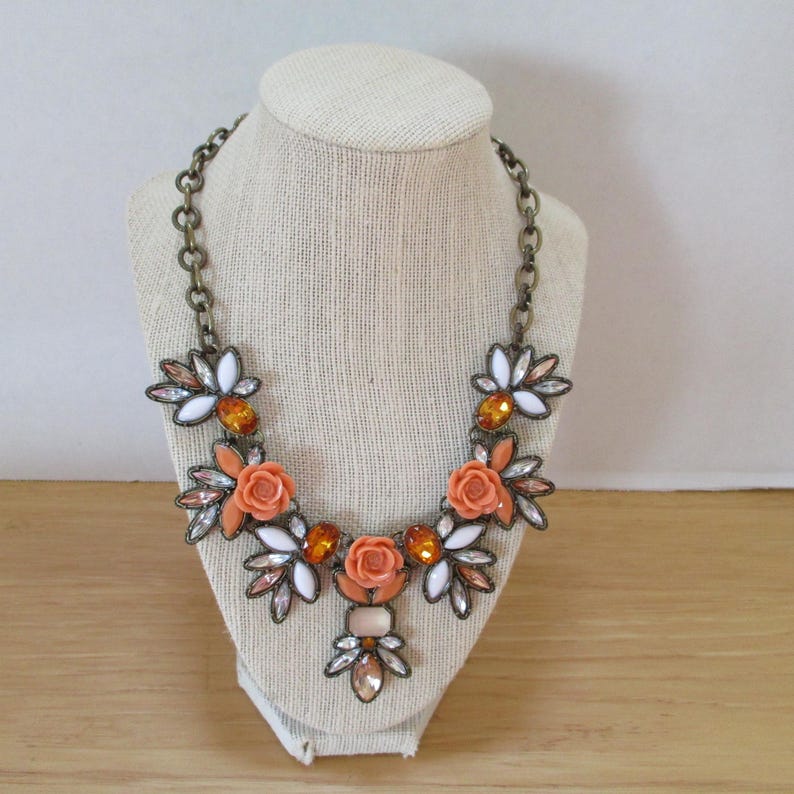 May include: A statement necklace with a floral design featuring orange and white flowers, clear and amber rhinestones, and a gold-tone chain.
