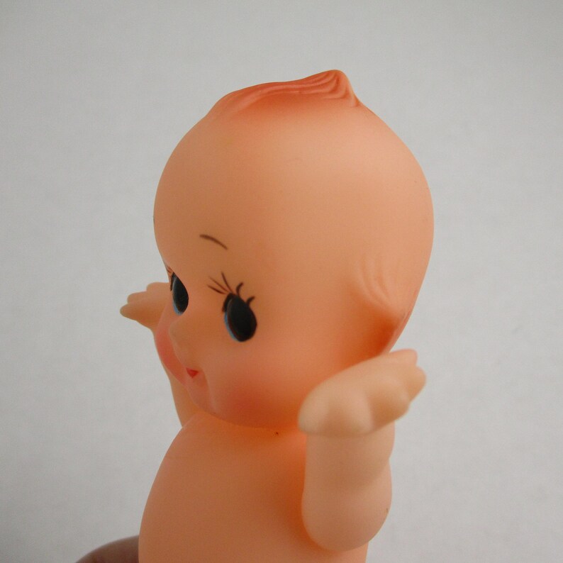 May include: A vintage-style baby doll with a peach-colored skin tone. The doll has large black eyes with long eyelashes, a small red mouth, and a tuft of reddish-orange hair. Its arms are raised, and it appears to be made of rubber or a similar material.