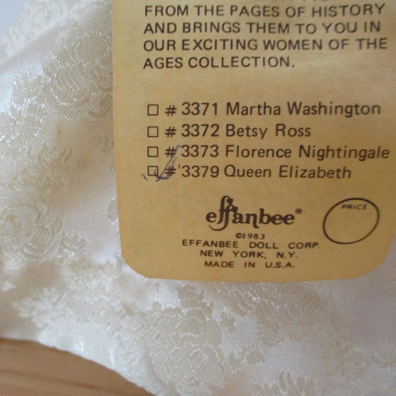 Puede incluir: Una etiqueta de papel marr&oacute;n con texto blanco que enumera los nombres de figuras hist&oacute;ricas, incluidas Martha Washington, Betsy Ross, Florence Nightingale y la reina Isabel. La etiqueta tambi&eacute;n incluye el texto "effanbee" y "&copy;1983 EFFANBEE DOLL CORP. NEW YORK, N.Y. MADE IN U.S.A."