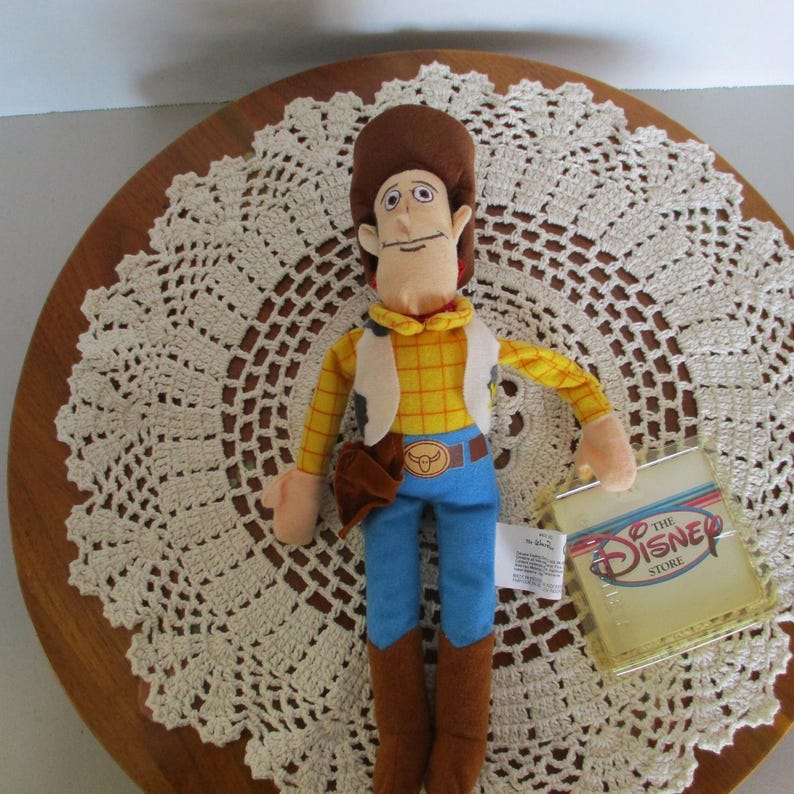 Puede incluir: Un peluche del personaje Woody de Toy Story de Disney. Woody viste una camisa a cuadros amarilla, pantalones azules, un chaleco blanco y botas marrones. Tiene un sombrero de vaquero marr&oacute;n y una funda marr&oacute;n. La etiqueta de Disney Store est&aacute; adjunta.
