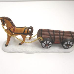 May include: A brown horse figurine with a black mane and harness, pulling a wooden cart loaded with logs. The horse and cart are connected by gold-colored chains and are mounted on a white, snow-textured base, creating a winter scene display.