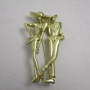 May include: Gold-toned metal brooch featuring a stylized couple, a woman in a hat and a man in a suit, standing side-by-side.