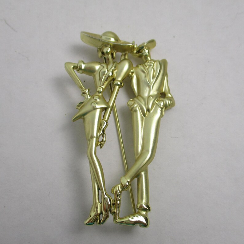 May include: Gold-toned metal brooch featuring a stylized couple, a woman in a hat and a man in a suit, standing side-by-side.