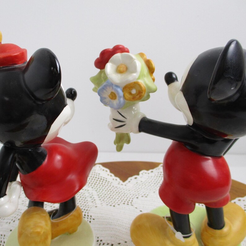 May include: Figurines of Mickey and Minnie Mouse. Mickey, in red shorts, offers a bouquet of flowers to Minnie, who wears a red dress and hat. Both figures have black ears and yellow shoes. The figurines are on a light green base.
