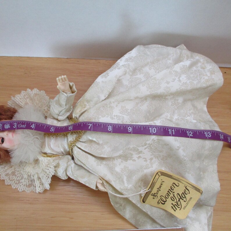 Puede incluir: Una mu&ntilde;eca de porcelana blanca que lleva un vestido blanco con ribetes dorados y un cuello de encaje blanco. La mu&ntilde;eca est&aacute; tumbada de espaldas con los brazos extendidos. La mu&ntilde;eca forma parte de la colecci&oacute;n Effanbee's Women of the Ages.