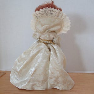 Puede incluir: Una mu&ntilde;eca de porcelana que lleva un vestido blanco con ribete de encaje y un cintur&oacute;n dorado. La mu&ntilde;eca tiene el pelo rojo y lleva una diadema de encaje blanco.