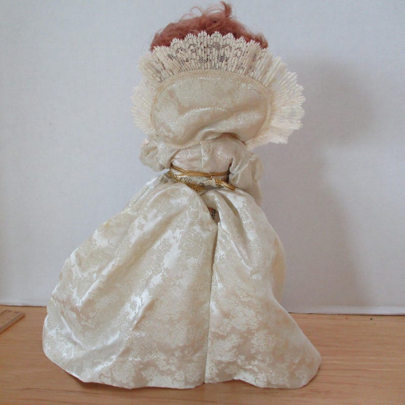 Puede incluir: Una mu&ntilde;eca de porcelana que lleva un vestido blanco con ribete de encaje y un cintur&oacute;n dorado. La mu&ntilde;eca tiene el pelo rojo y lleva una diadema de encaje blanco.