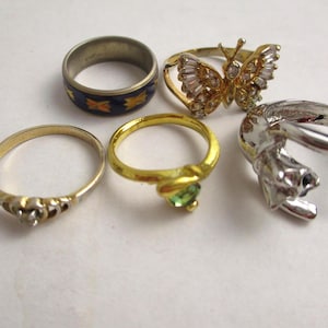 May include: A collection of five rings in various styles. One ring features a blue band with gold starburst designs. Another is a gold ring with a butterfly design, and a third has a green gemstone. The rings are set against a white background.