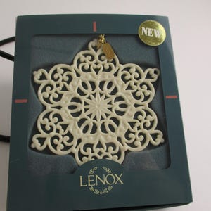 May include: Ivory-colored snowflake ornament in a dark teal box. The ornament has an intricate, openwork design with a central starburst pattern. The box has the brand name "LENOX" and a gold "NEW" sticker.