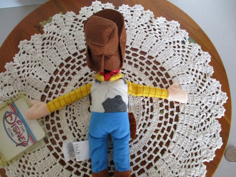Puede incluir: Mu&ntilde;eco de peluche de Woody, con sombrero de vaquero marr&oacute;n, camisa a cuadros amarilla, vaqueros azules y chaleco con estampado de vaca. La mu&ntilde;eca es de Disney Store y est&aacute; sobre un tapete de ganchillo blanco.