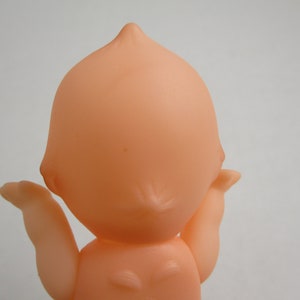 May include: A vintage-style, peach-colored baby doll, viewed from the back. The doll has a rounded head with a small point at the top and outstretched arms. The doll's body is smooth and simple, with a classic design.