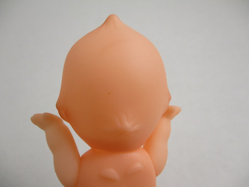 May include: A vintage-style, peach-colored baby doll, viewed from the back. The doll has a rounded head with a small point at the top and outstretched arms. The doll's body is smooth and simple, with a classic design.