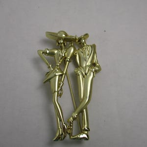 May include: Gold-toned metal brooch featuring a stylized couple, a woman in a hat and a man in a suit, standing side-by-side.