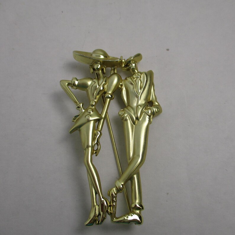 May include: Gold-toned metal brooch featuring a stylized couple, a woman in a hat and a man in a suit, standing side-by-side.