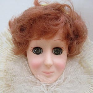 Puede incluir: Una cabeza de mu&ntilde;eca de porcelana con cabello rojo rizado, ojos verdes y un cuello de encaje blanco.