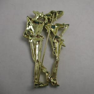 May include: Gold-toned metal brooch featuring two stylized figures, one holding a staff, with intricate details and a textured finish.