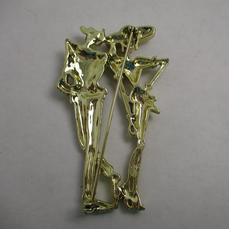 May include: Gold-toned metal brooch featuring two stylized figures, one holding a staff, with intricate details and a textured finish.
